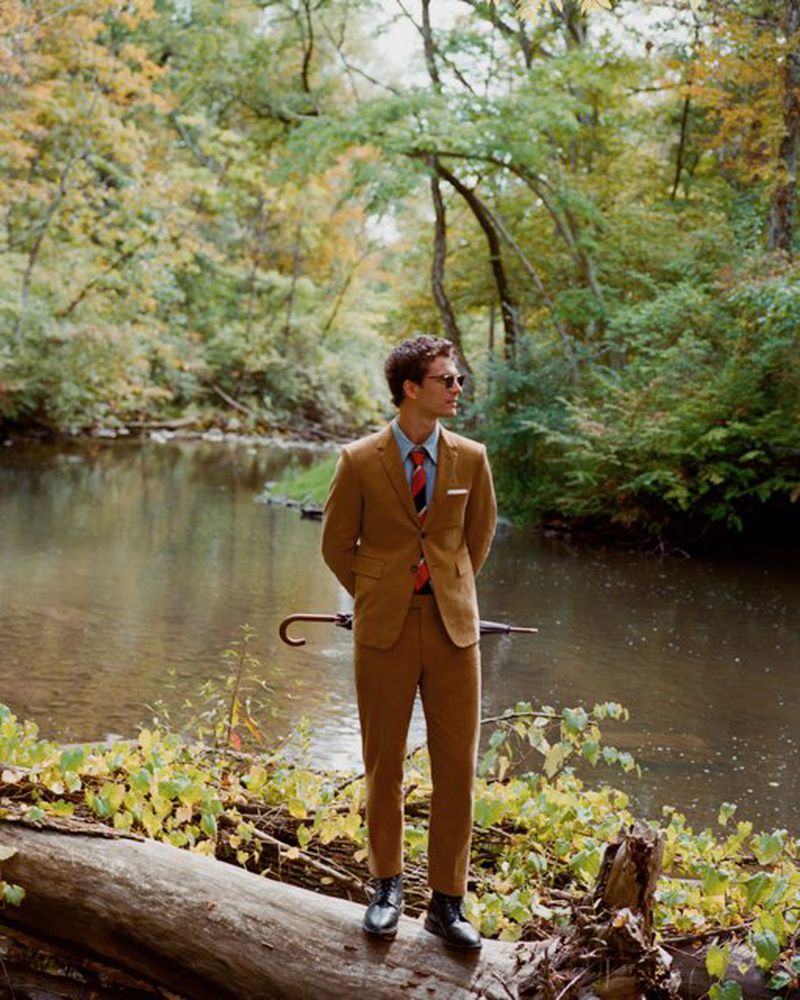 Adult male in tan suit stands on a log near river in autumn woods, wearing sunglasses, with umbrella propped behind him.