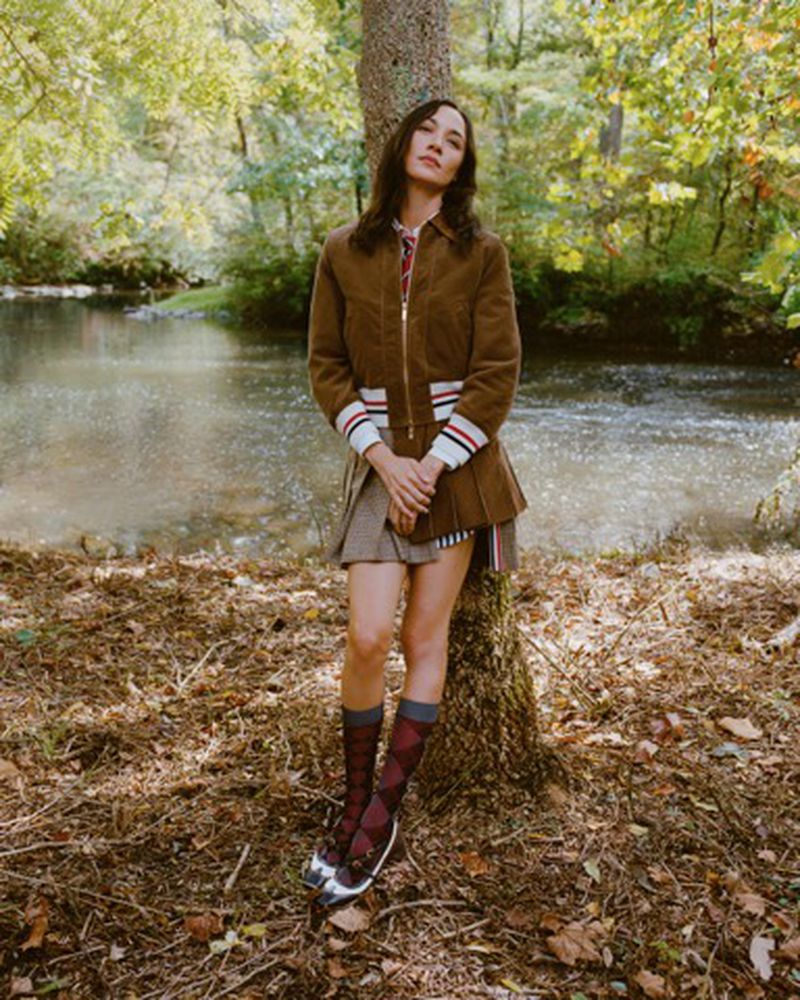 Woman poses in a brown jacket, pleated skirt, and knee socks near a creek. Outdoors, nature scene with water and trees.