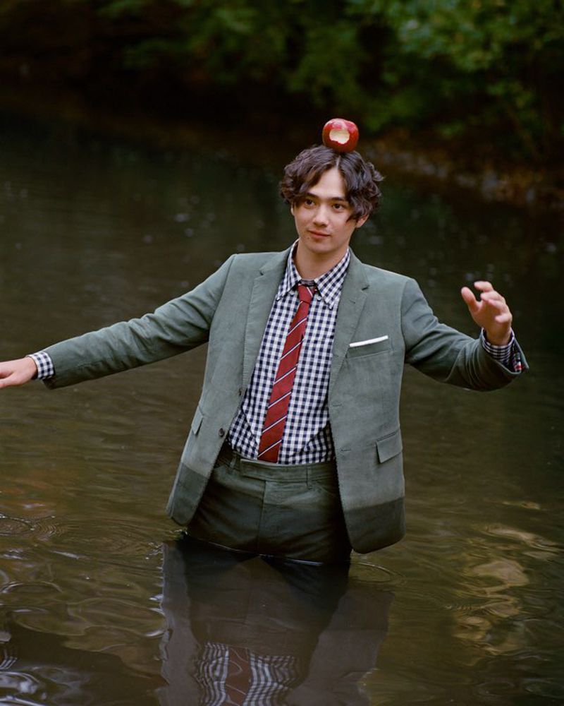 A young man in a green suit stands waist-deep in a river with an apple balanced on his head in an outdoor setting.