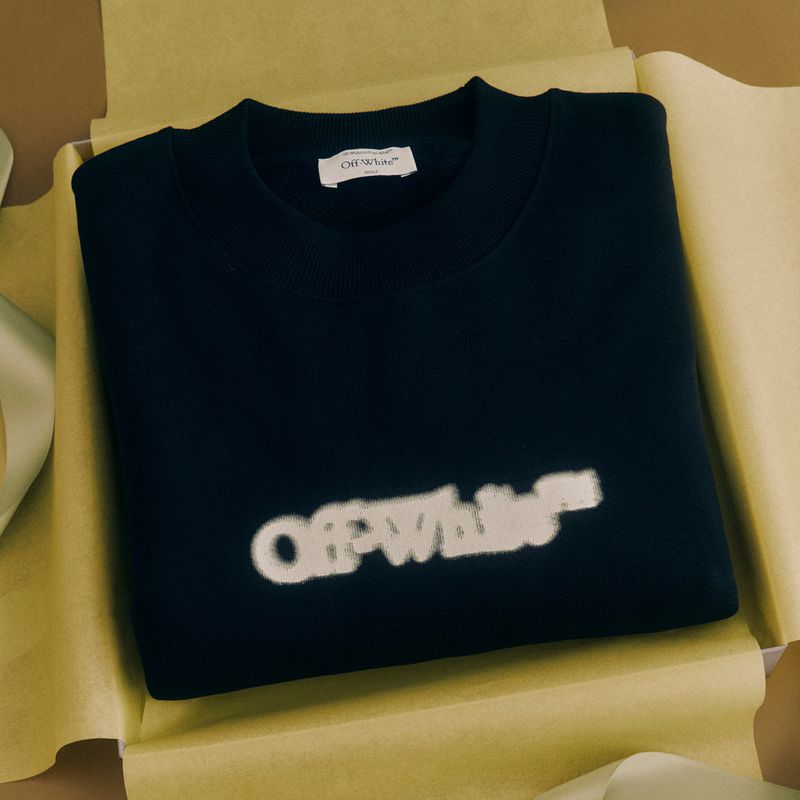 Navy sweater with white Off-White logo in a yellow box, presented with a light green ribbon, clean studio shot, neutral backdrop.