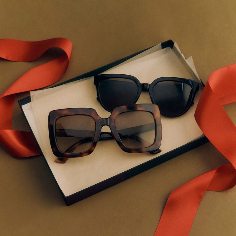 Two pairs of sunglasses, one black and one tortoiseshell, are displayed in a box with a red ribbon against a gold background.