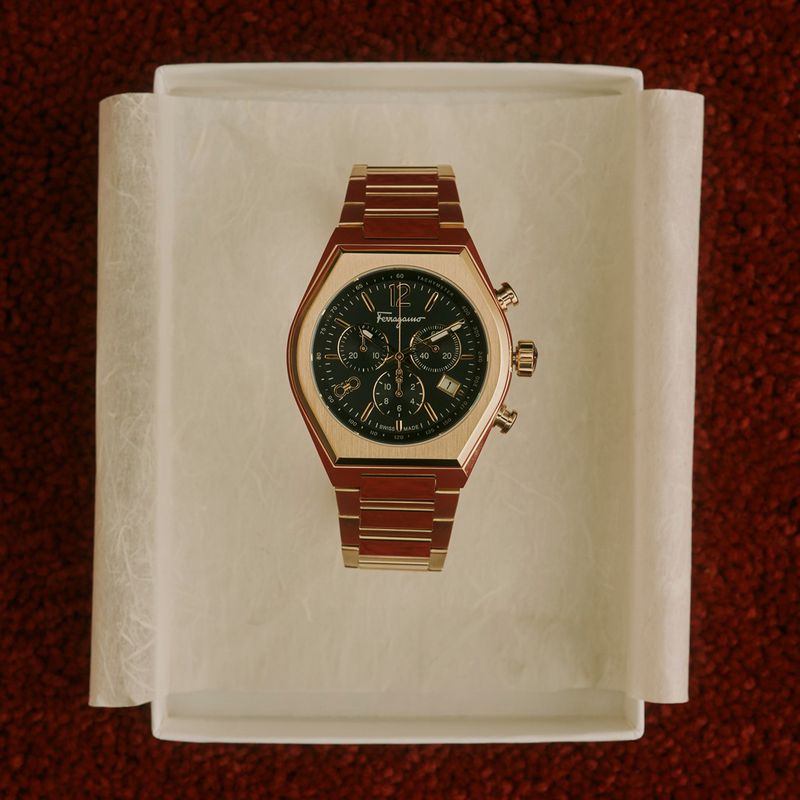 Elegant rose-gold Salvatore Ferragamo chronograph wristwatch on a textured cream-colored platform against a red speckled background.