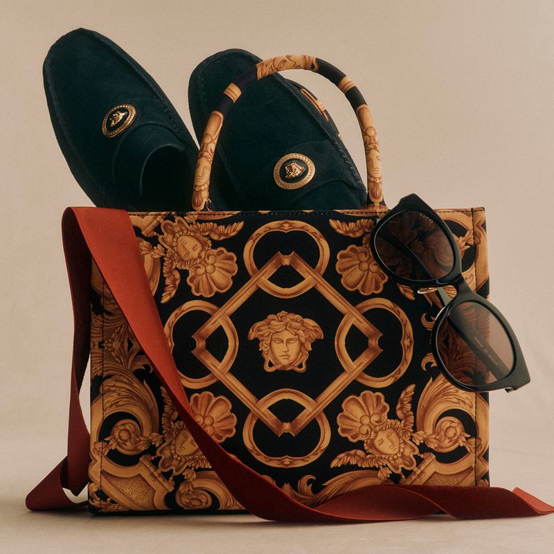 Fashion still life featuring a patterned Versace handbag, dark loafers with gold accents, sunglasses and a red-orange ribbon.