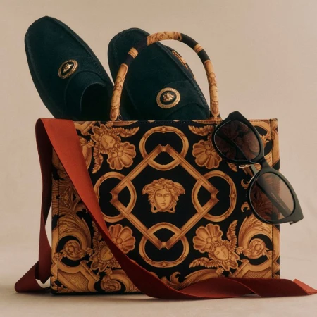 Fashion still life featuring a patterned Versace handbag, dark loafers with gold accents, sunglasses and a red-orange ribbon.