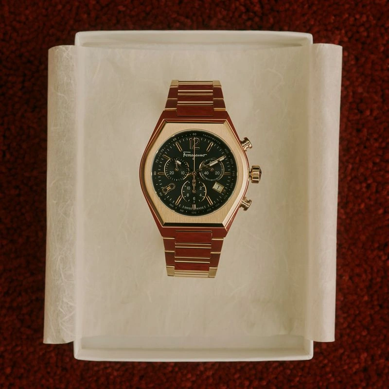 Gold-tone chronograph watch with a black dial displayed inside an open white gift box lined with tissue paper.