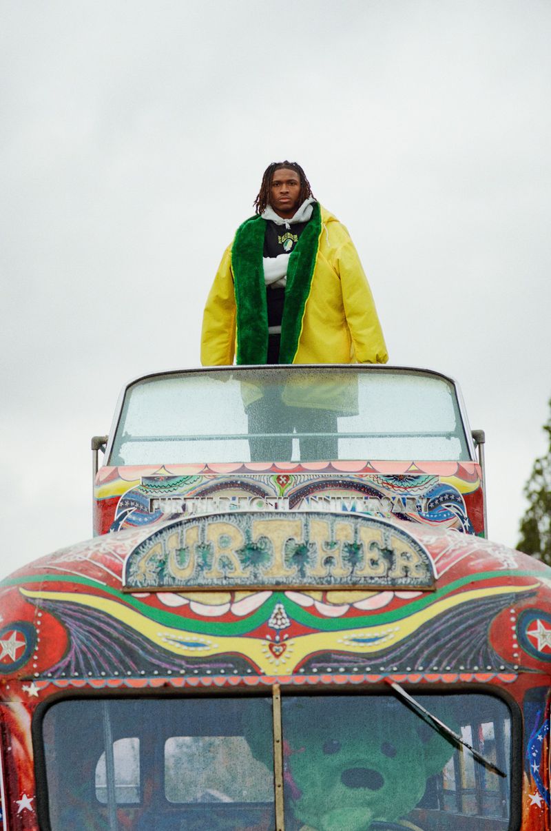 Adult male in yellow jacket standing on top of colorfully painted vehicle with a green stuffed animal in the front window against cloudy sky.