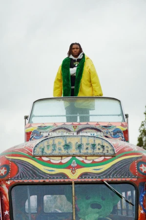 Adult male in yellow jacket standing on top of colorfully painted vehicle with a green stuffed animal in the front window against cloudy sky.