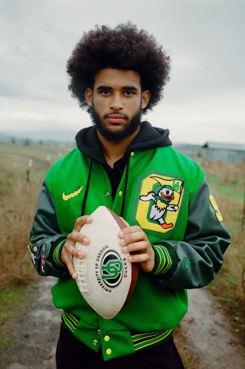 A young man with afro hairstyle holds a football and poses in a green jacket with the University of Oregon logo outdoors.