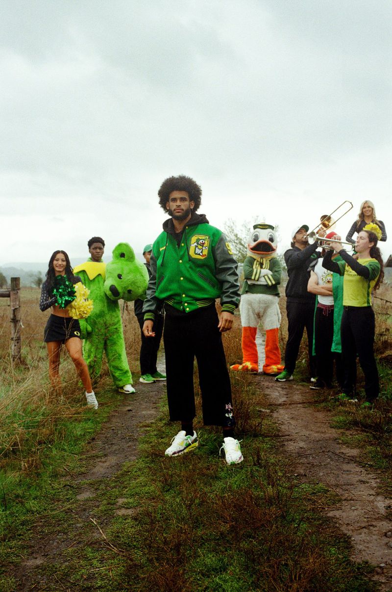 Group shot of a person in a letterman jacket, cheerleaders with pom-poms, mascots, and a marching band on a grassy path outdoors.