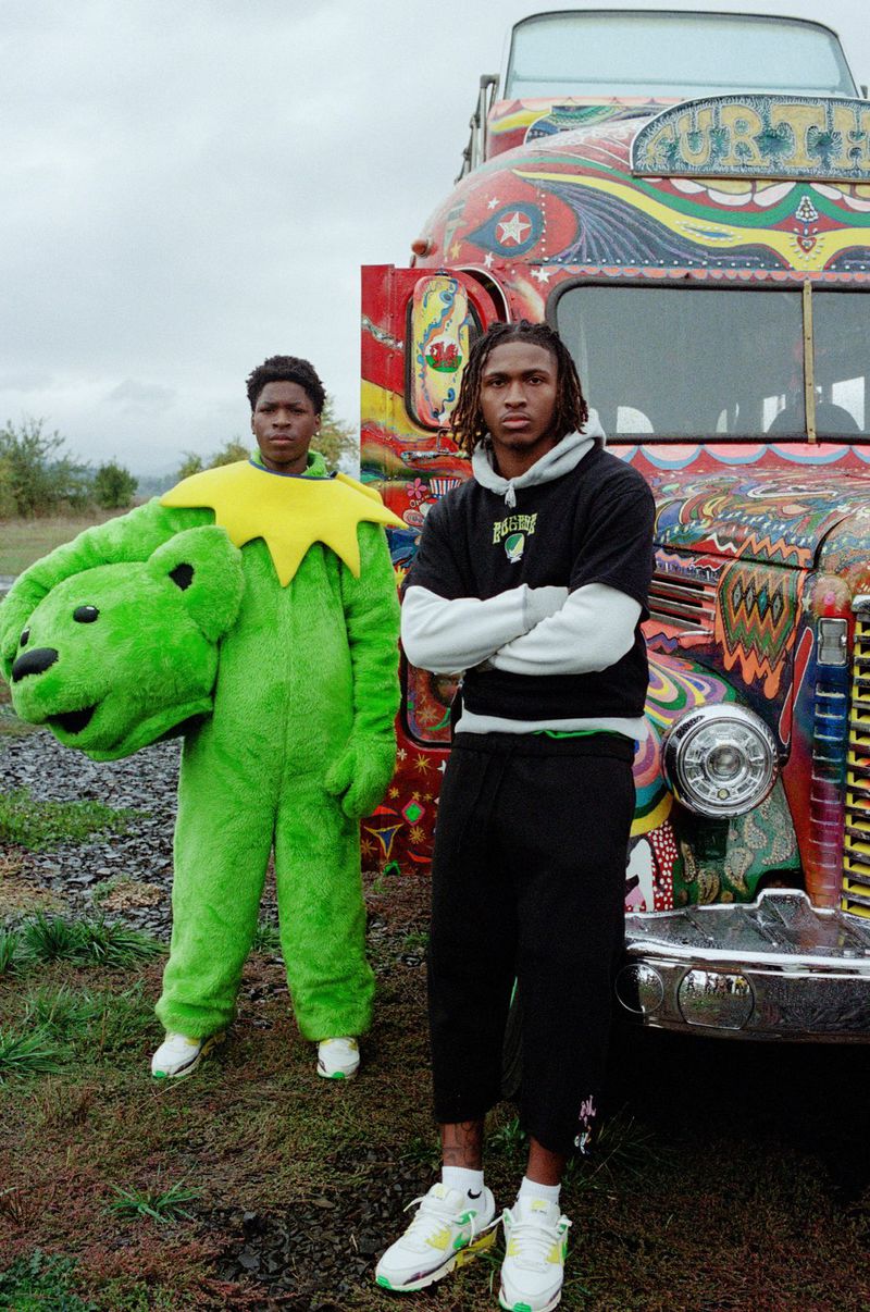 Two people pose outdoors in front of a painted bus. One person wears a Grateful Dead bear costume, while the other wears streetwear.