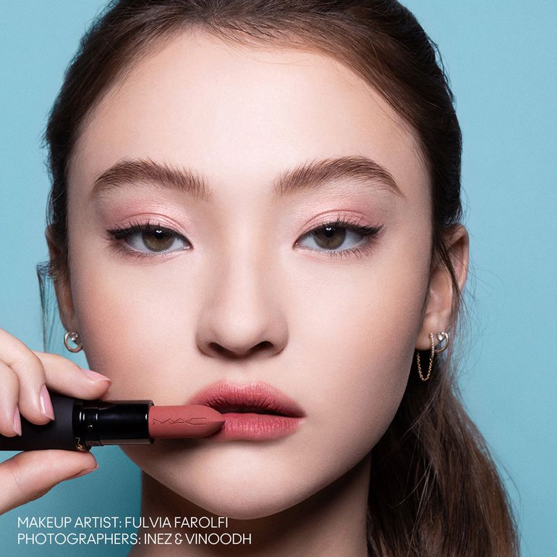 Close-up beauty shot: young woman with light skin, brown hair, wearing pink eyeshadow and lipstick, holding lipstick tube against a blue background.