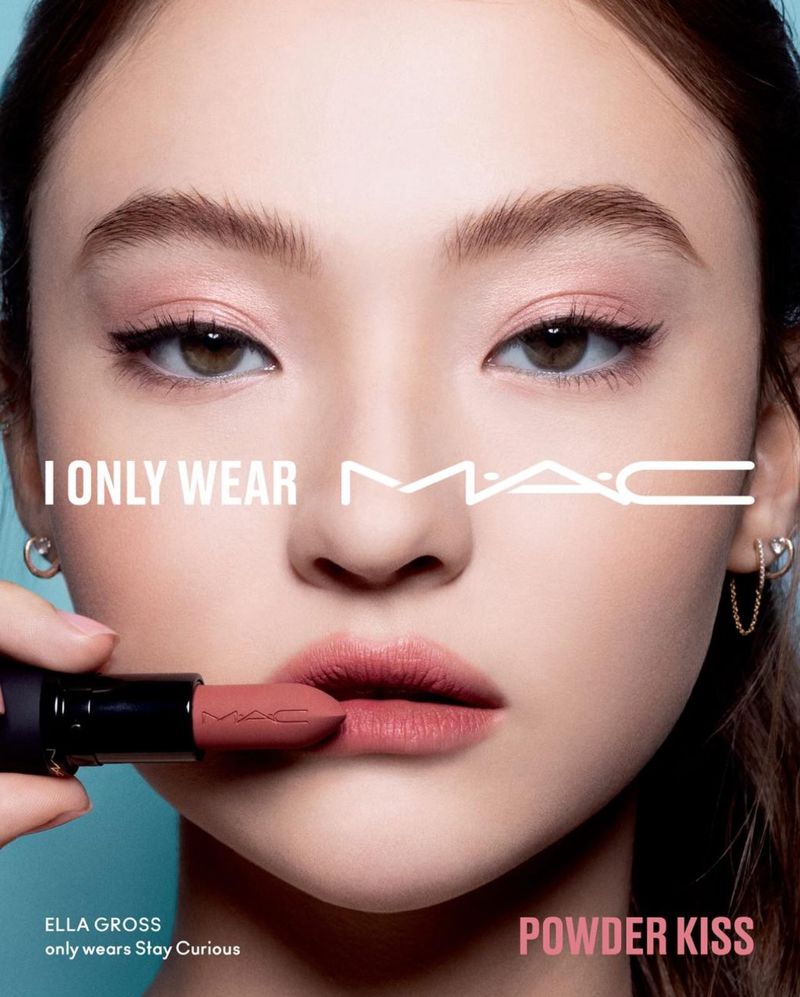 Close-up of a woman's face with light skin, wearing pink eyeshadow and lipstick, holding a MAC lipstick tube in front of her mouth.