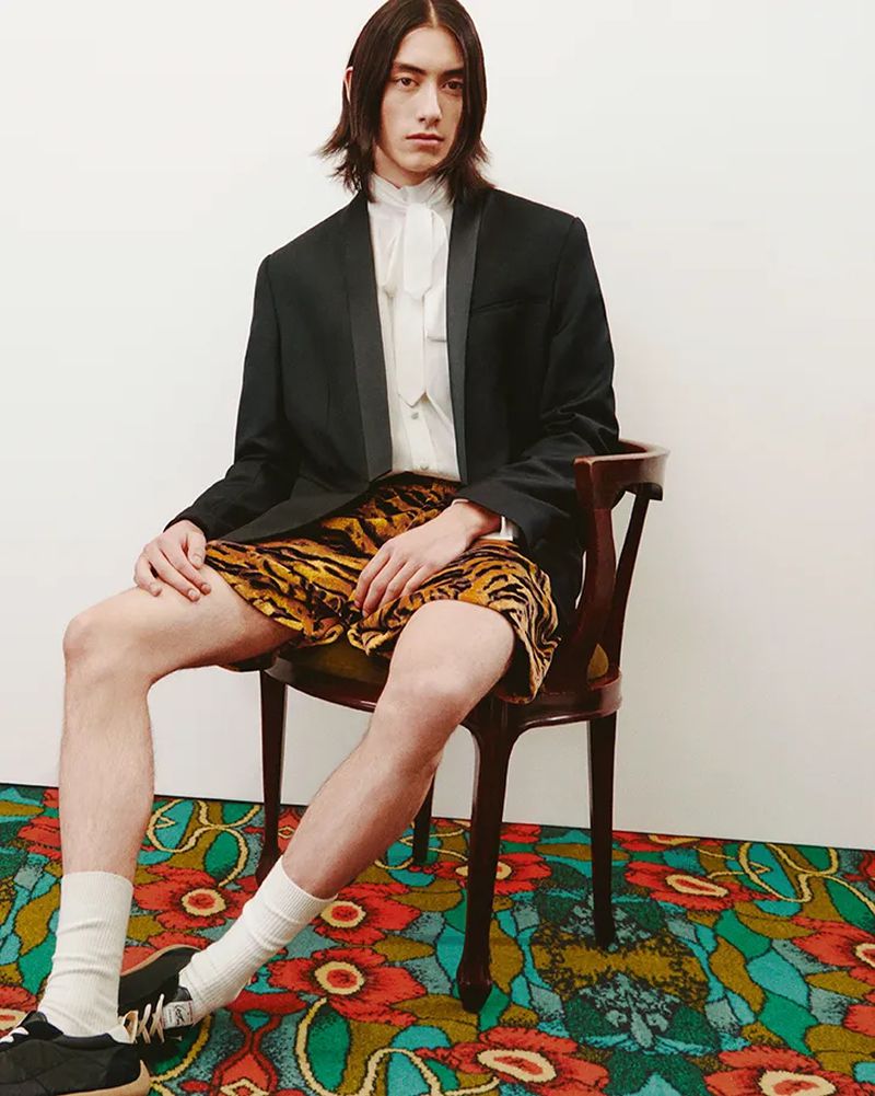 Model seated on a wooden chair wearing a black blazer, white shirt, patterned shorts, and socks on a floral carpet