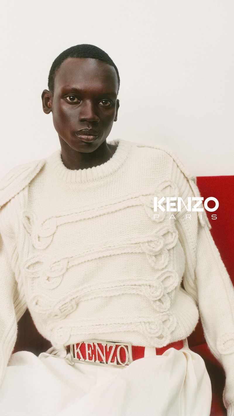 Headshot of a Black male model with close-cropped hair, wearing a textured knit sweater and a logo belt against a white background.