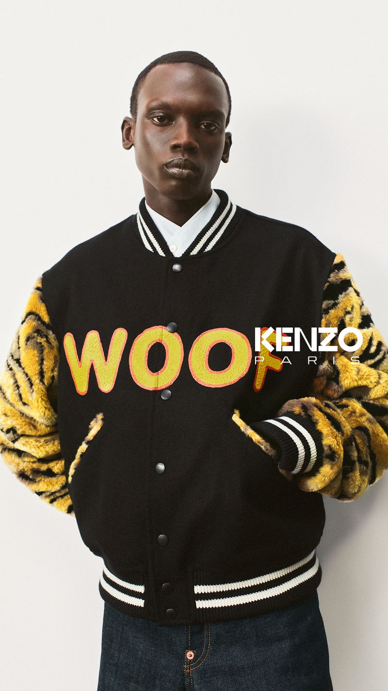 Male fashion model in a studio shot, wearing a letterman jacket with faux fur sleeves, and jeans, against a neutral background.