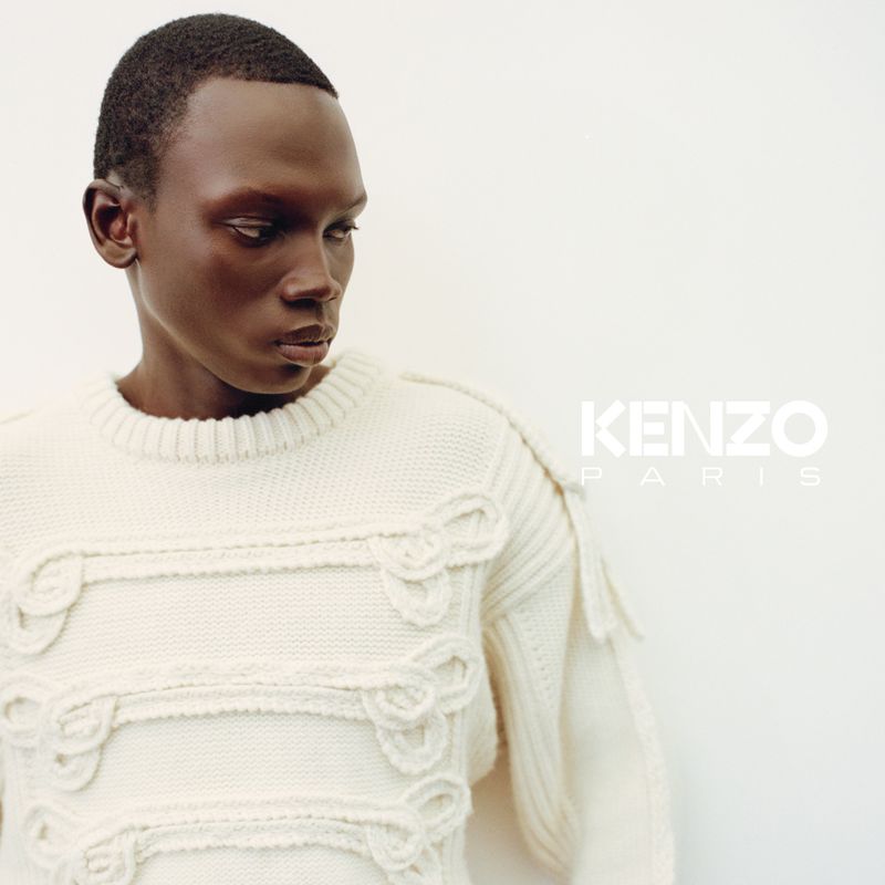 Portrait of a Black man wearing a cream-colored sweater. The KENZO logo is visible to the right of the model on a neutral-colored background.