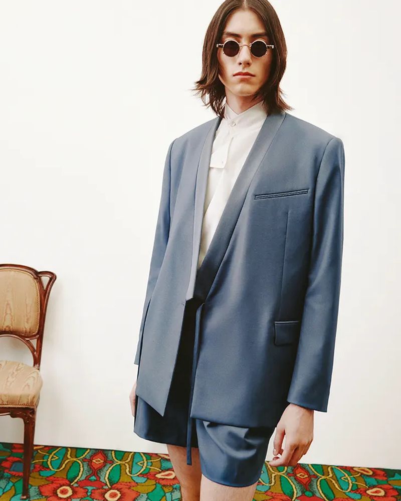 A person in a blue suit with shorts and round sunglasses stands in front of a white wall with an antique chair and patterned floor.