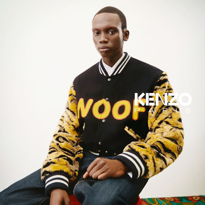 A person wearing a varsity jacket with tiger-striped sleeves is sitting indoors, photographed in front of a neutral background with Kenzo Paris logo.