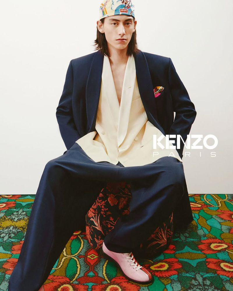 Man in oversized navy suit with printed headscarf sits on a patterned floral ottoman, posing in a fashion studio against a white backdrop.