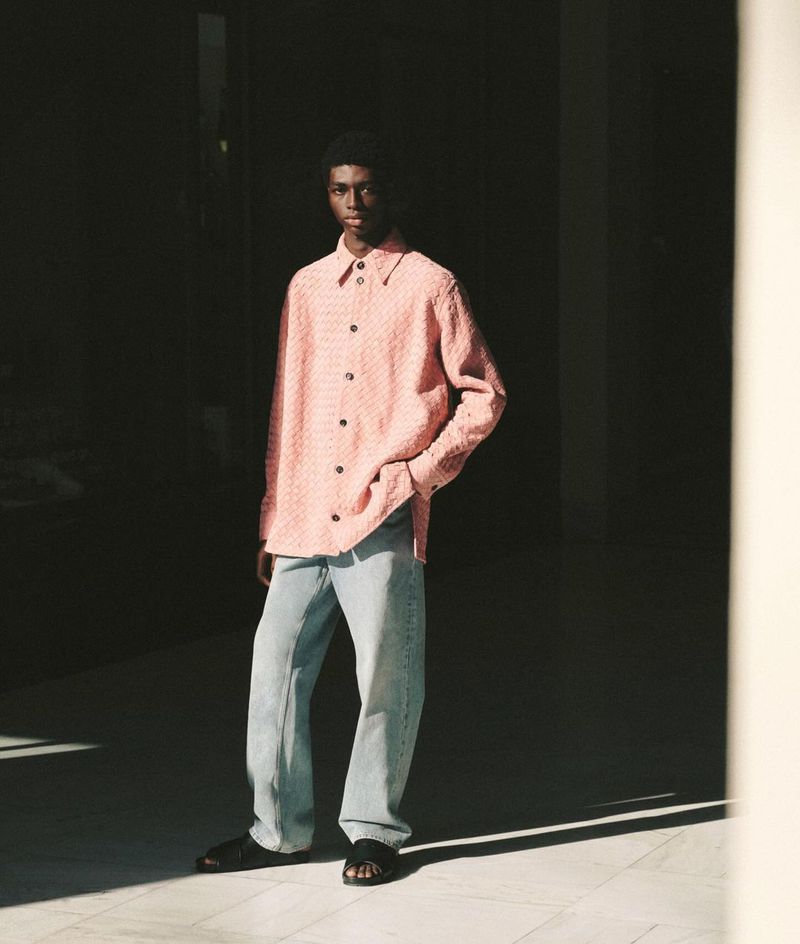 Portrait of a Black male model in pink shirt and blue jeans, standing indoors with a hand in his pocket, contrasted by shadows.