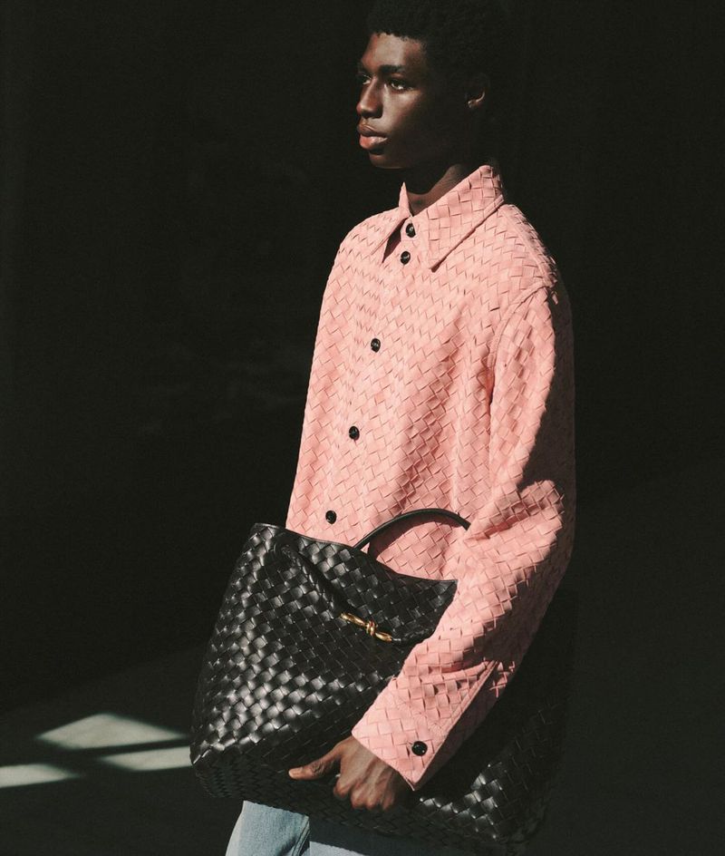 Black model carrying a woven black leather bag, wearing a pink woven button-down shirt and blue jeans, indoor fashion shot.