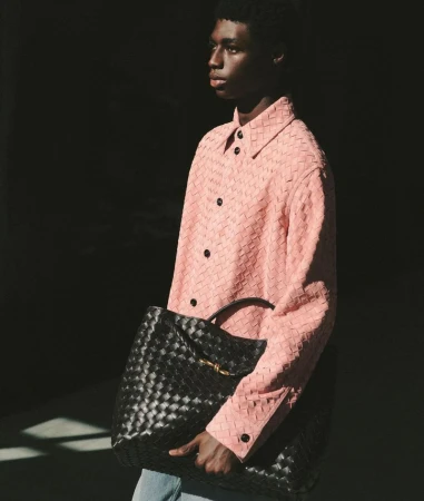 Black model carrying a woven black leather bag, wearing a pink woven button-down shirt and blue jeans, indoor fashion shot.