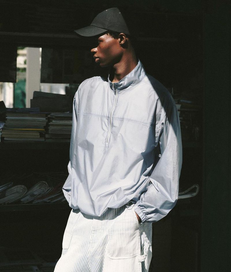 Young man in a black cap and gray quarter-zip pullover poses in front of a shelf full of newspapers in an urban setting with natural light.