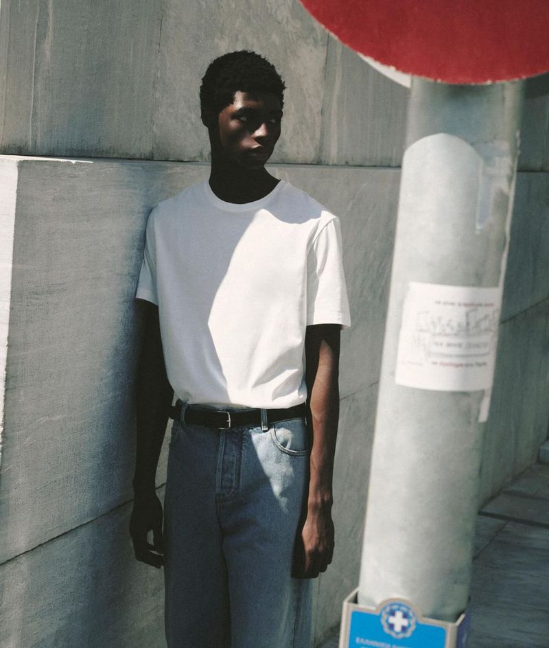 A young Black male model poses outdoors in a white t-shirt and jeans against a concrete wall and a gray pole with red circular object.