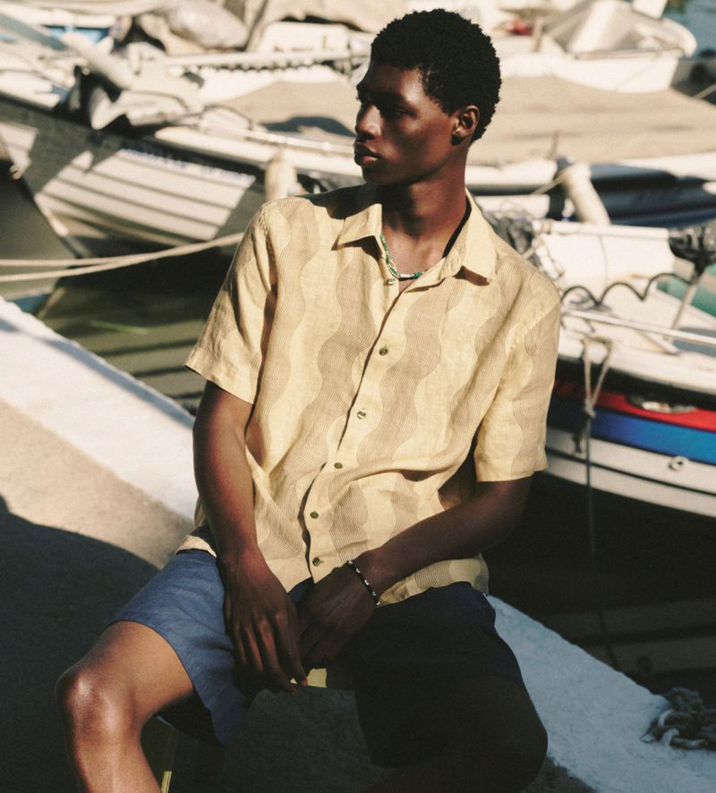 A young black male model is sitting outdoors by the water wearing a beige patterned short-sleeved shirt and dark shorts on a sunny day.