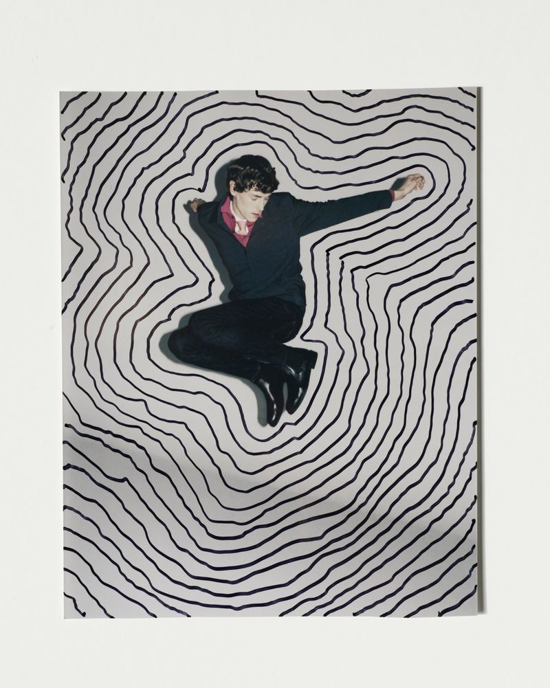 A man in mid-air wearing a sweater and dress pants poses in front of a hand-drawn black and white topographic pattern background.