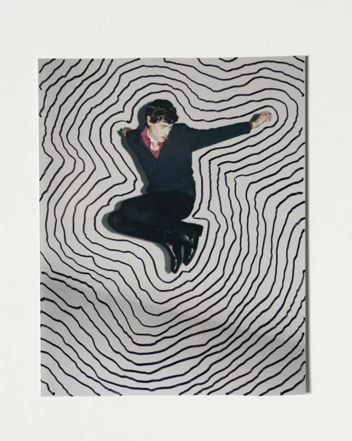 A man in mid-air wearing a sweater and dress pants poses in front of a hand-drawn black and white topographic pattern background.