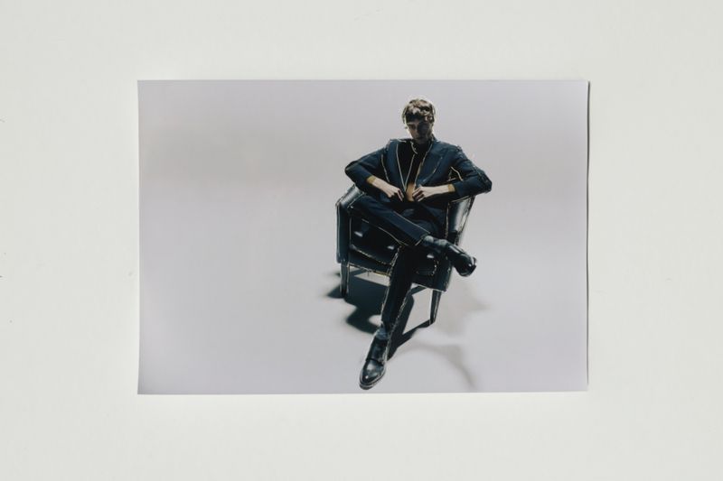 A male fashion model sits in a leather chair indoors wearing a dark suit and boots against a solid grey background in a portrait photograph.