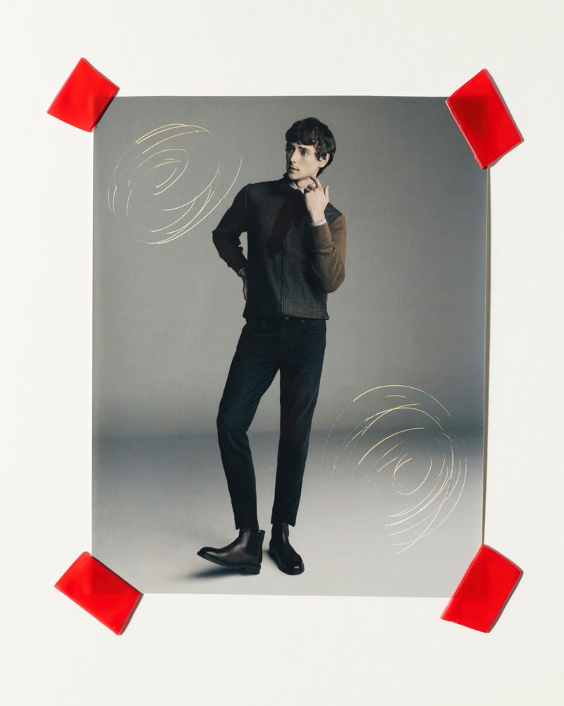 Studio shot of a male fashion model in a dark sweater, black pants, and boots, posing against a gray backdrop with red tape accents.