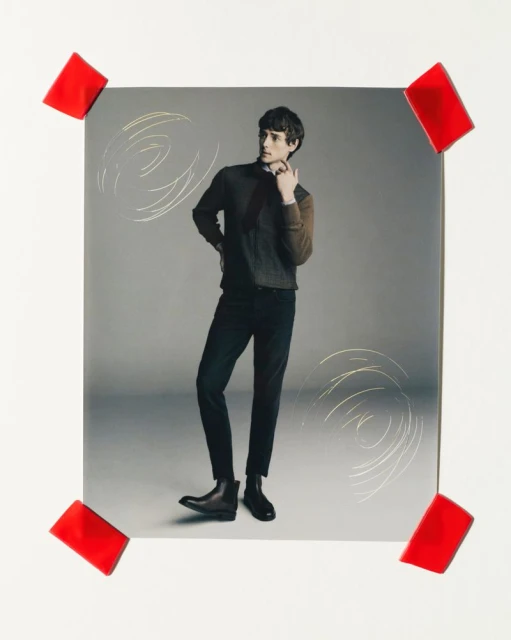 Studio shot of a male fashion model in a dark sweater, black pants, and boots, posing against a gray backdrop with red tape accents.