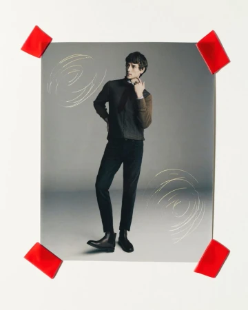 Studio shot of a male fashion model in a dark sweater, black pants, and boots, posing against a gray backdrop with red tape accents.