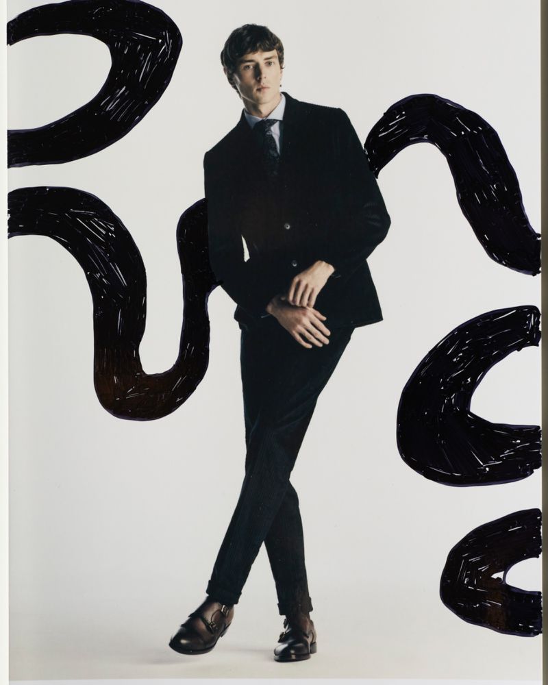 A stylish young man in a black suit strikes a pose in a studio setting against a backdrop of bold, abstract black strokes.