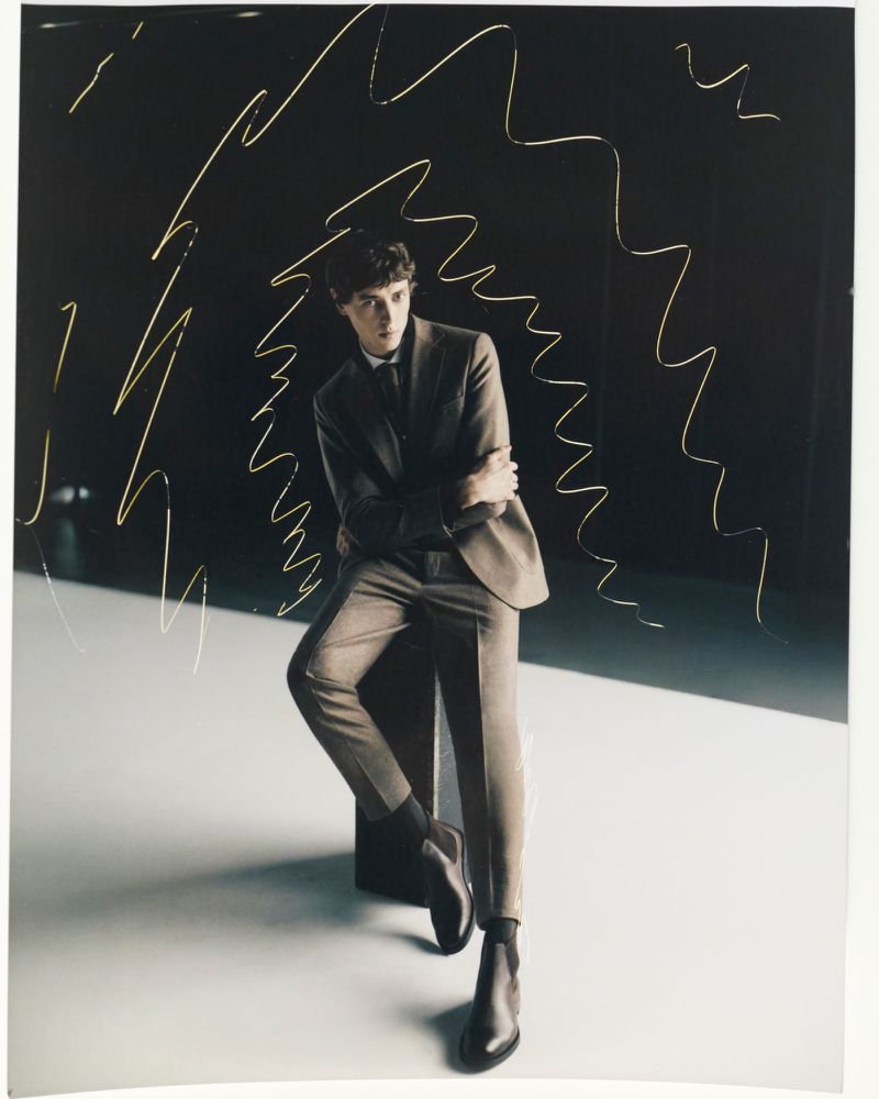 Man in suit leans against dark backdrop with gold lines. Indoor portrait showcasing fashion and posing with neutral expression.
