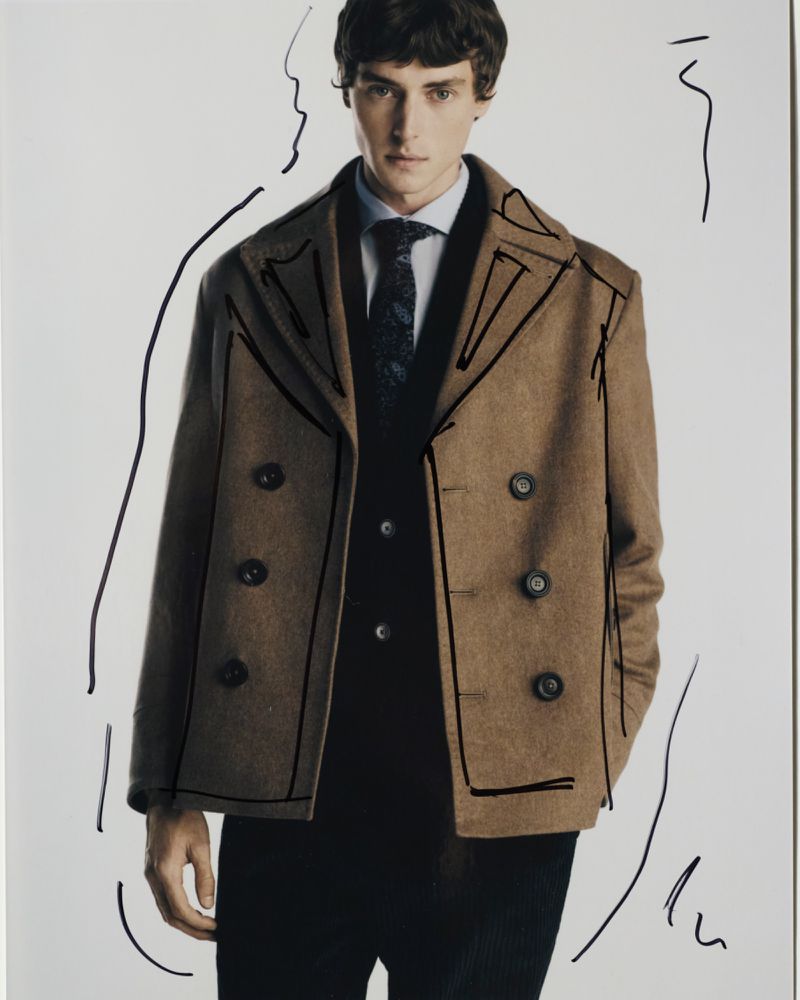 Portrait of a young adult male wearing a tan double-breasted coat over a dark vest, collared shirt, and tie, with black graphic lines.