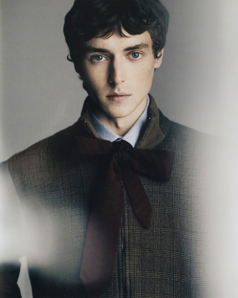 Close-up studio portrait of a young adult male with dark curly hair, wearing a plaid jacket and burgundy bow, looking at the camera.
