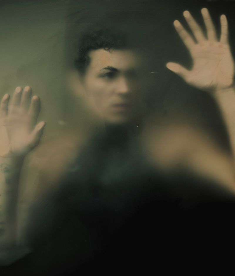 Portrait of a man with curly hair and tattoos behind frosted glass, raising both hands; the light is diffused and the atmosphere is soft.