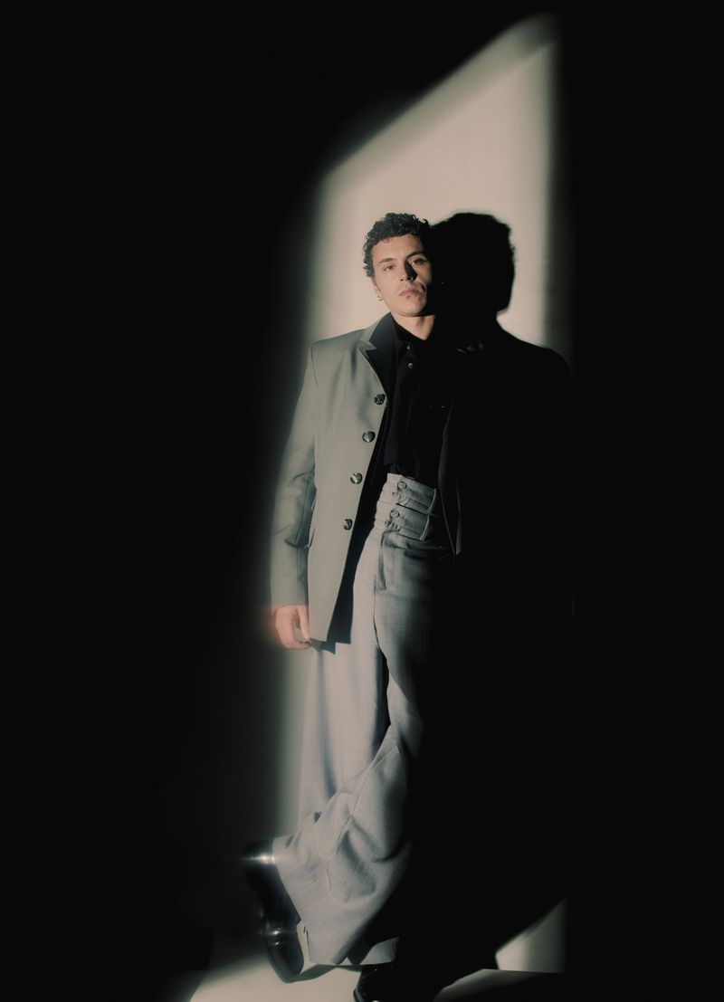 Man in a grey suit poses in dramatic lighting, creating a strong shadow. He is wearing black shirt and trousers, with a confident expression.