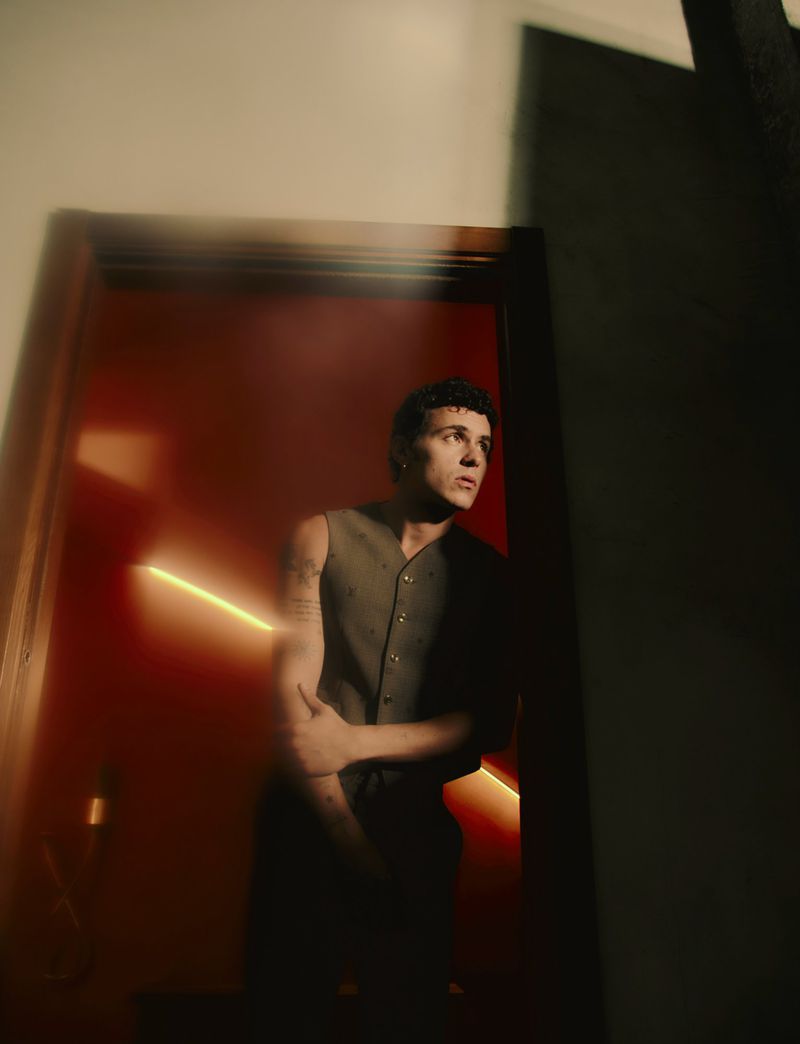 Portrait of a young adult male in a dark vest standing in a doorway with a red wall and neon light.