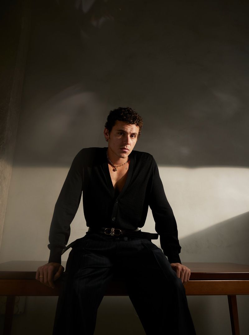 Portrait of a young adult male wearing a black cardigan and pants, sitting on a wooden table in a dimly lit indoor setting.