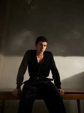 Portrait of a young adult male wearing a black cardigan and pants, sitting on a wooden table in a dimly lit indoor setting.