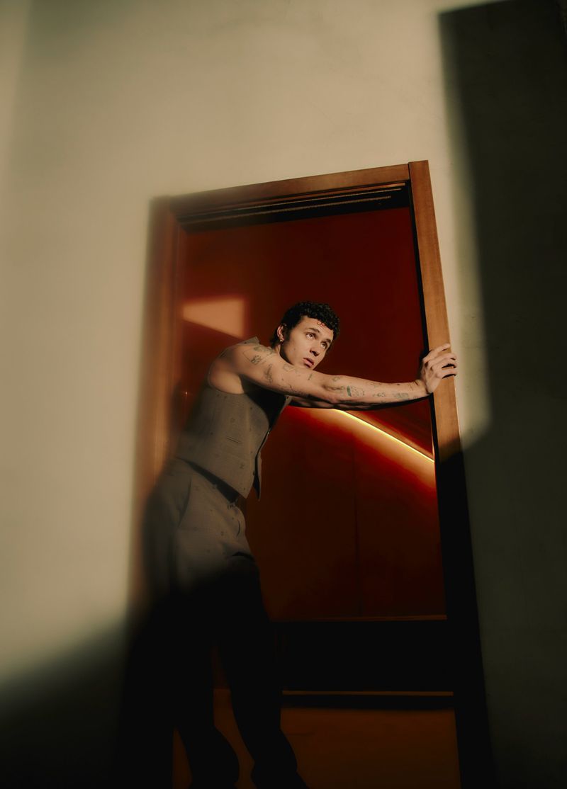 Adult male with tattoos leaning against an open doorway with a red-orange glow in a sleeveless top and suit pants in a dimly lit interior.