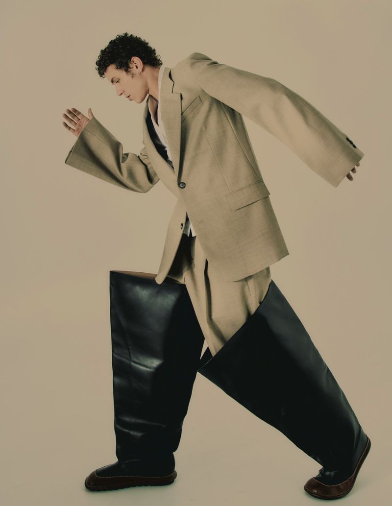 Man in a tan suit poses in studio, wearing large black boots with a geometric shape, captured with studio lighting on a neutral background.