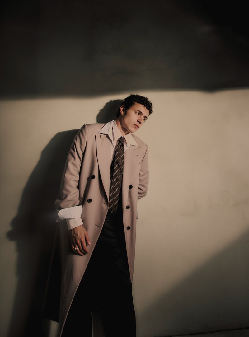 Man in tan overcoat and tie in a studio setting with directional lighting, creating shadows on the neutral wall, posing.