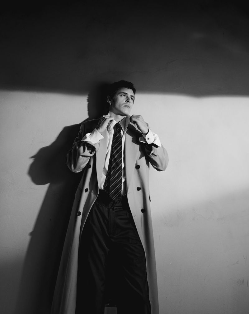 Black and white portrait of a man adjusting his shirt collar while wearing a long trench coat and tie indoors against a wall.