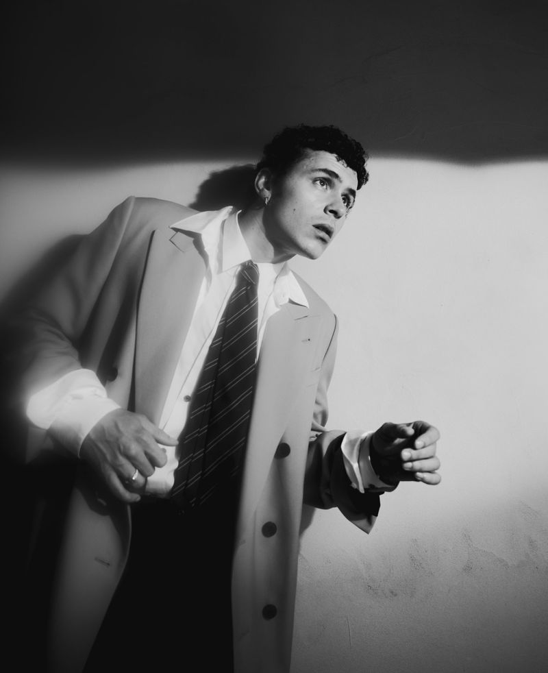 Black and white portrait of a young adult man in a suit jacket and tie, looking up and away, posing in an indoor photo studio.