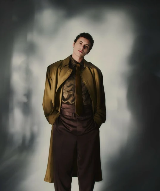 Stylish young man in olive trench coat, brown shirt and pants, standing in studio with soft lighting and diffused background.